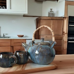 Könnte beinhalten: Eine blaue und braune Teekanne aus Keramik mit einem Bambusgriff, zwei passende Tassen und ein Holzbrett. Die Teekanne und die Tassen haben eine gesprenkelte Glasur.