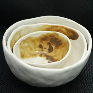 May include: Three white ceramic bowls nested together. The bowls have a textured, uneven surface and are glazed with a light brown colour. The inside of the bowls have a darker brown stain.