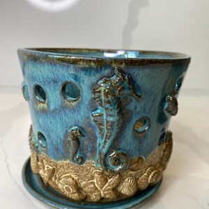 May include: A ceramic planter with a turquoise glaze and brown accents. The planter features seahorse and seashell relief details. It has a matching saucer and several circular openings around the top.