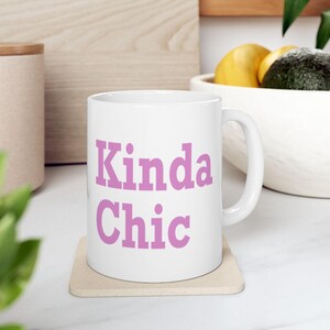 May include: A white ceramic mug with a handle, featuring the text Kinda Chic printed in a bold, light purple serif font on the front. The mug is positioned on a square, light-coloured coaster, displaying a clean and simple design.