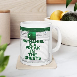 May include: White coffee mug featuring a green and white design of a spreadsheet with the text "NATHANIEL IS A FREAK IN THE SHEETS".