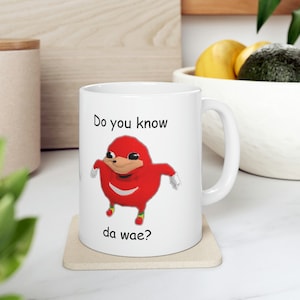 Puede incluir: Taza de cerámica blanca con un personaje de dibujos animados rojo y el texto "Do you know da wae?" El personaje tiene un cuerpo rojo, una cara beige y guantes blancos. La taza tiene un asa blanca y está sobre un posavasos beige.