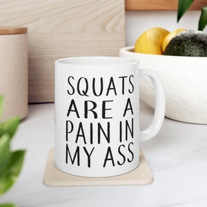May include: White ceramic coffee mug with black text that reads "SQUATS ARE A PAIN IN MY ASS".