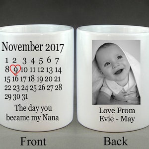 Op de afbeelding: Witte keramische mok met een zwart-witfoto van een baby op de achterkant. De voorkant van de mok heeft een kalender voor november 2017 met de 9e gemarkeerd en een rood hart. De tekst "De dag dat je mijn Nana werd" staat onder de kalender.