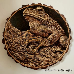 May include: A detailed wood-burned art piece featuring a frog perched on a branch. The artwork is circular, with a natural wood edge. The frog is brown with intricate details, and the branch has textured lines. The text "islandarts&crafts" is at the bottom.
