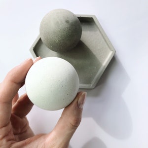 K&ouml;nnte beinhalten: Eine hellgr&uuml;ne Kugel wird in einer Hand gehalten, mit einer passenden Kugel und einer sechseckigen Schale im Hintergrund. Die Objekte bestehen aus Beton und haben eine strukturierte Oberfl&auml;che. Die Farbpalette ist neutral mit Gr&uuml;n- und Graut&ouml;nen.