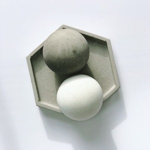 K&ouml;nnte beinhalten: Eine sechseckige Betonschale h&auml;lt zwei kugelf&ouml;rmige Objekte. Eine Kugel ist dunkelgrau, die andere ist heller, cremewei&szlig;. Die Schale und die Kugeln befinden sich auf wei&szlig;em Hintergrund. Die Objekte scheinen dekorativ zu sein.