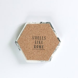 Puede incluir: Posavasos hexagonal con centro de corcho y borde con dise&ntilde;o de m&aacute;rmol. El centro de corcho presenta el texto "Smells Like Home" en letras negras. El posavasos est&aacute; dise&ntilde;ado para bebidas.