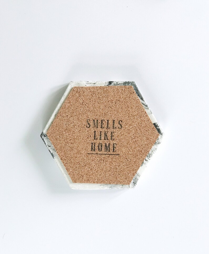 Puede incluir: Posavasos hexagonal con centro de corcho y borde con dise&ntilde;o de m&aacute;rmol. El centro de corcho presenta el texto "Smells Like Home" en letras negras. El posavasos est&aacute; dise&ntilde;ado para bebidas.