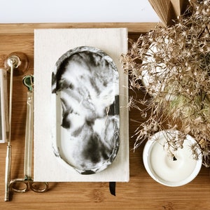 Pu&ograve; includere: Un vassoio decorativo con un motivo marmorizzato bianco e nero &egrave; appoggiato su un vassoio di legno, accanto a una candela, un taccuino e accessori per candele color oro. Fiori secchi e un ventaglio di carta completano la composizione. Il vassoio &egrave; lungo circa 15 cm.