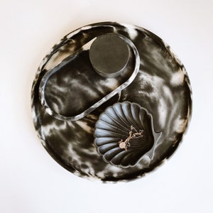 Pu&ograve; includere: Un vassoio decorativo con un motivo marmorizzato bianco e nero. Il vassoio contiene un piccolo oggetto circolare nero, un oggetto ovale trasparente e una ciotola a forma di conchiglia. Una collana d'oro con un ciondolo &egrave; all'interno della ciotola.