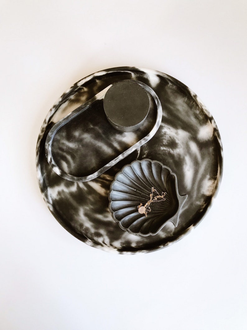 Pu&ograve; includere: Un vassoio decorativo con un motivo marmorizzato bianco e nero. Il vassoio contiene un piccolo oggetto circolare nero, un oggetto ovale trasparente e una ciotola a forma di conchiglia. Una collana d'oro con un ciondolo &egrave; all'interno della ciotola.