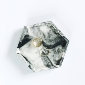Puede incluir: Una bandeja hexagonal con un dise&ntilde;o veteado en blanco y negro contiene dos anillos. Un anillo es dorado y el otro tiene una banda plateada con una piedra transparente. La bandeja es un art&iacute;culo decorativo para guardar joyas.