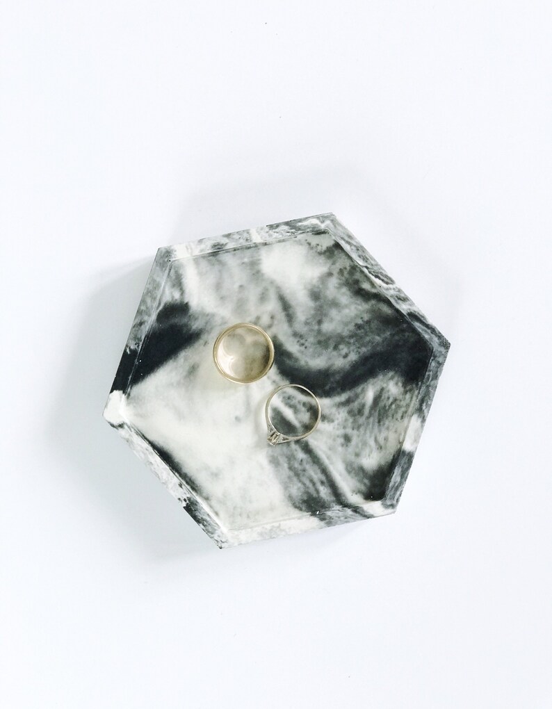 Puede incluir: Una bandeja hexagonal con un dise&ntilde;o veteado en blanco y negro contiene dos anillos. Un anillo es dorado y el otro tiene una banda plateada con una piedra transparente. La bandeja es un art&iacute;culo decorativo para guardar joyas.