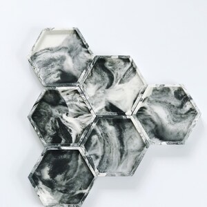 Puede incluir: Un conjunto de siete posavasos hexagonales con un dise&ntilde;o veteado en blanco y negro. Los posavasos est&aacute;n dispuestos en un grupo, mostrando los patrones arremolinados &uacute;nicos del material. Los bordes son de color gris s&oacute;lido.