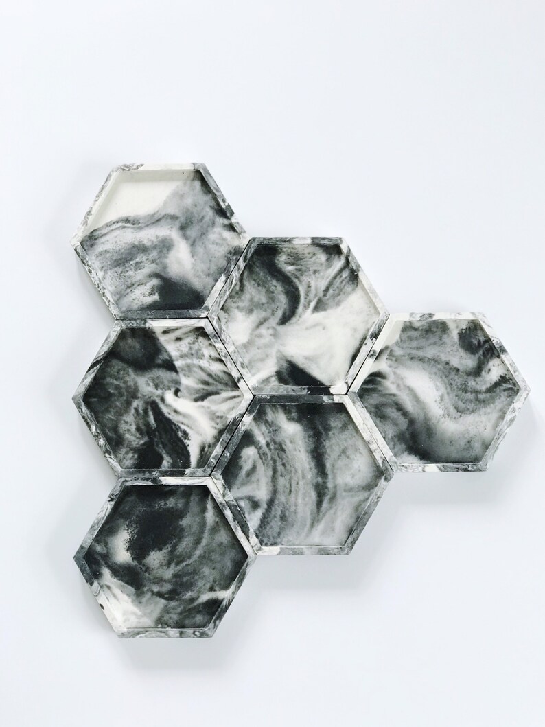 Puede incluir: Un conjunto de siete posavasos hexagonales con un dise&ntilde;o veteado en blanco y negro. Los posavasos est&aacute;n dispuestos en un grupo, mostrando los patrones arremolinados &uacute;nicos del material. Los bordes son de color gris s&oacute;lido.