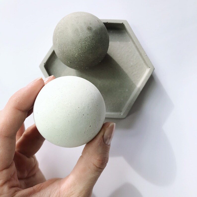 K&ouml;nnte beinhalten: Eine hellgr&uuml;ne Kugel wird in einer Hand gehalten, mit einer passenden Kugel und einer sechseckigen Schale im Hintergrund. Die Objekte bestehen aus Beton und haben eine strukturierte Oberfl&auml;che. Die Farbpalette ist neutral mit Gr&uuml;n- und Graut&ouml;nen.