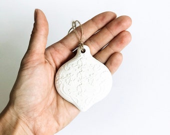 May include: A white, teardrop-shaped ornament with an embossed floral design, suspended by a natural twine loop. The ornament is held in a hand against a white background. The design is a classic holiday decoration.