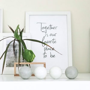 K&ouml;nnte beinhalten: Ein wei&szlig; gerahmter Druck mit dem Text "Together is our favorite place to be" steht auf einem Regal mit dekorativen Kugeln. Eine kleine Pflanze in einem Glasr&ouml;hrchen und einem Holzrahmen f&uuml;gt einen Hauch von Natur hinzu.