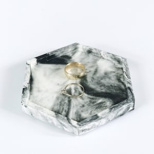 Puede incluir: Una bandeja hexagonal con un dise&ntilde;o veteado en blanco y negro. Dos anillos de oro est&aacute;n colocados en la bandeja. Uno es una banda simple, y el otro tiene un peque&ntilde;o diamante. La bandeja est&aacute; sobre un fondo blanco.