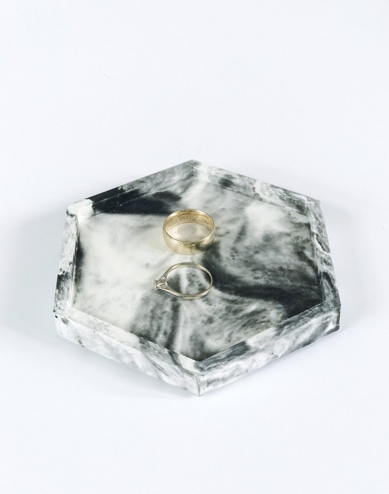 Puede incluir: Una bandeja hexagonal con un dise&ntilde;o veteado en blanco y negro. Dos anillos de oro est&aacute;n colocados en la bandeja. Uno es una banda simple, y el otro tiene un peque&ntilde;o diamante. La bandeja est&aacute; sobre un fondo blanco.