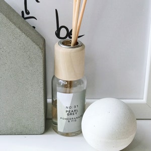 K&ouml;nnte beinhalten: Eine transparente Glas-Diffusorflasche mit Holzverschluss und St&auml;bchen, beschriftet mit "No:31 Pearl Grey Pomegranate & Fig". Eine wei&szlig;e Kugel und ein graues Betonobjekt in Hausform sind ebenfalls vorhanden.