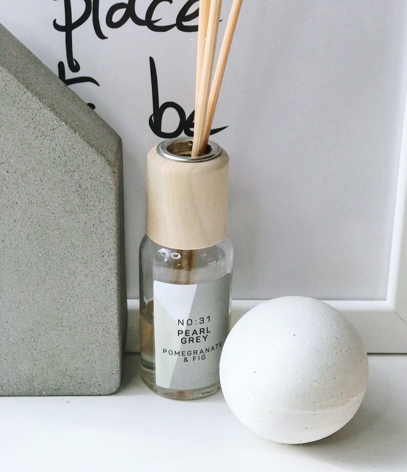 K&ouml;nnte beinhalten: Eine transparente Glas-Diffusorflasche mit Holzverschluss und St&auml;bchen, beschriftet mit "No:31 Pearl Grey Pomegranate & Fig". Eine wei&szlig;e Kugel und ein graues Betonobjekt in Hausform sind ebenfalls vorhanden.