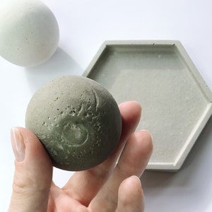 K&ouml;nnte beinhalten: Eine Sammlung von Beton-Wohnaccessoires. Das Bild zeigt eine graue Kugel in einer Hand, eine hellgraue Kugel und ein sechseckiges Tablett. Die Kugel hat eine strukturierte Oberfl&auml;che mit einem geschnitzten Design. Die Artikel sind auf wei&szlig;em Hintergrund.