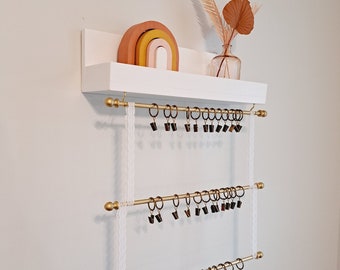 May include: White shelf with three gold rods and black clips. The shelf has a rainbow-colored wooden toy and a vase with dried flowers on it.