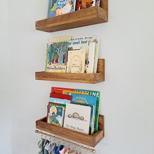 May include: Three wooden shelves with a gold metal rod underneath the bottom shelf. The shelves are filled with children's books. The books have colorful covers with illustrations and text. The rod has several colorful fabric loops hanging from it.