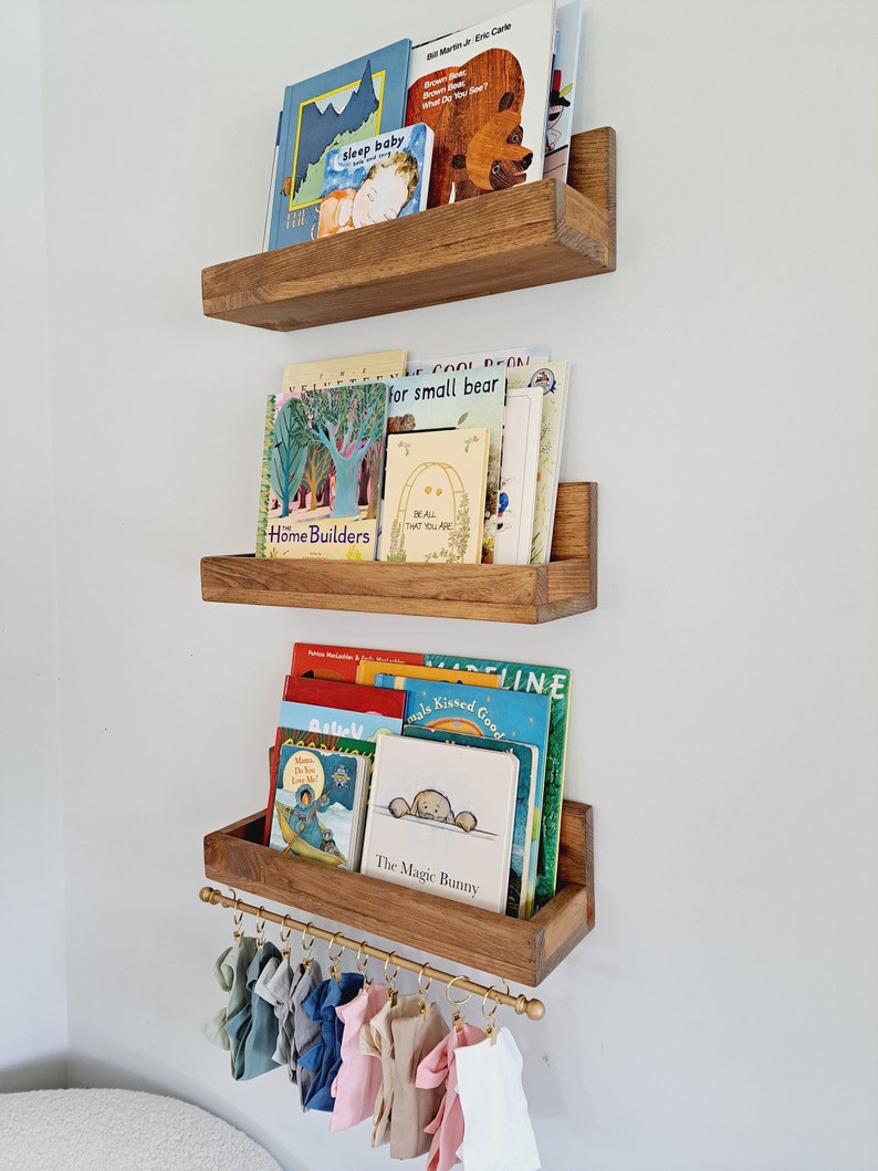 May include: Three wooden shelves with a gold metal rod underneath the bottom shelf. The shelves are filled with children's books. The books have colorful covers with illustrations and text. The rod has several colorful fabric loops hanging from it.