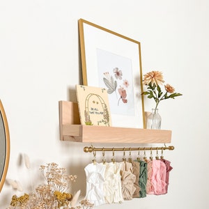 May include: A wooden shelf with a gold metal rod and clips for hanging items. The shelf has a book with the text "Be all that you are" and a framed print with watercolor flowers. There are several fabric headbands hanging from the rod.
