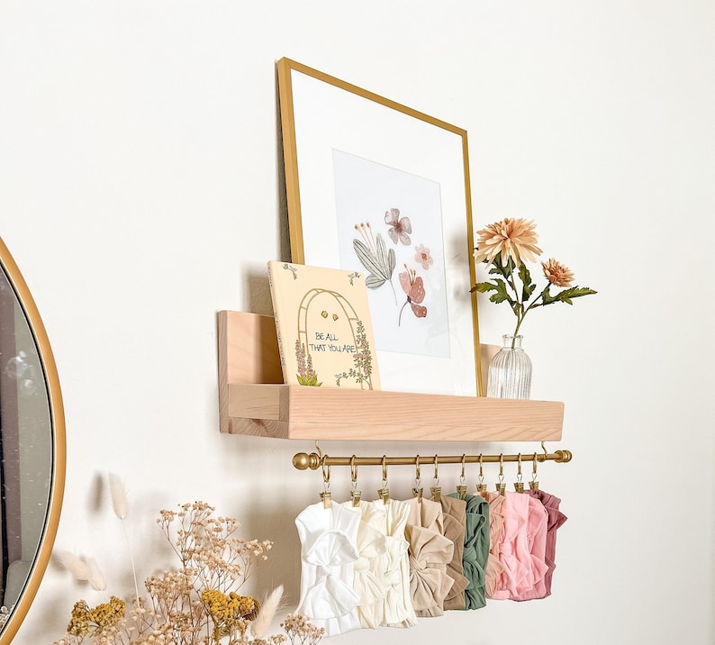 May include: A wooden shelf with a gold metal rod and clips for hanging items. The shelf has a book with the text "Be all that you are" and a framed print with watercolor flowers. There are several fabric headbands hanging from the rod.