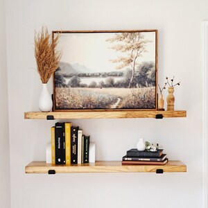 May include: Two wooden shelves with black metal brackets, holding a framed landscape painting, books, and decorative vases. The painting features a field with a lake and trees in the background. The books are stacked on the lower shelf, and the vases are on the upper shelf.