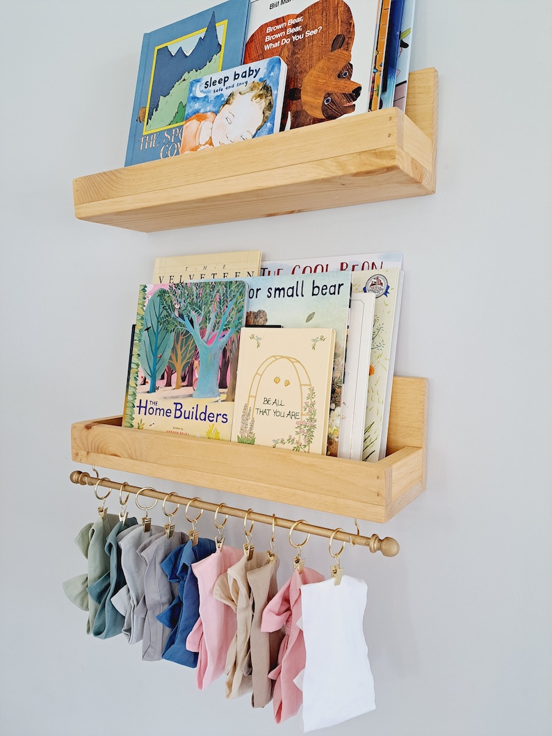 May include: Two wooden shelves with a gold metal rod underneath. The shelves are filled with children's books. The rod has several colorful fabric headbands hanging from it.