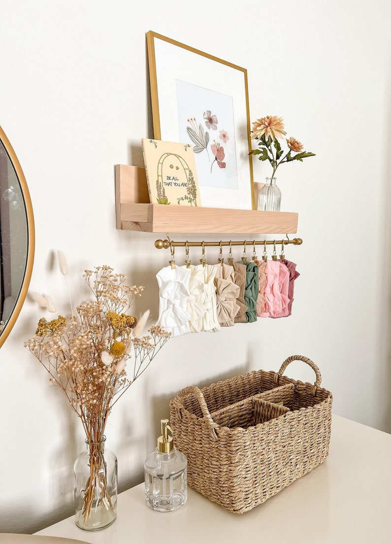 May include: A wooden shelf with a gold metal rod and clips holding a variety of fabric headbands in white, brown, green, and pink. A small book with the text "Be all that you are" is on the shelf. A framed print with watercolor flowers is above the shelf. A vase with dried flowers and a clear glass soap dispenser are on a table below the shelf.