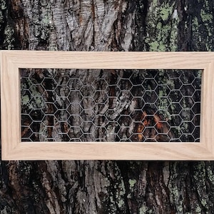 May include: A rectangular wooden frame with a chicken wire mesh insert. The frame is made of light-colored wood and is set against a textured tree bark background. The wire mesh has a hexagonal pattern.