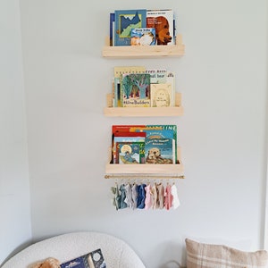 May include: Three wooden shelves with books on them, including "Sleep Baby", "The Story of Babar", "The Velveteen Rabbit", "Cool Room", "Small Bear", "Home Builders", "Sally", "I Love My Mama Because", and "One Snowy Night". The shelves are mounted on a white wall. Below the shelves is a gold metal rod with colorful fabric scraps hanging from it.