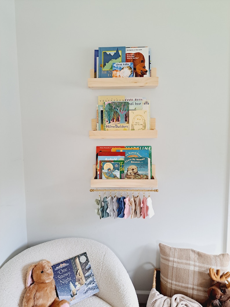 May include: Three wooden shelves with books on them, including "Sleep Baby", "The Story of Babar", "The Velveteen Rabbit", "Cool Room", "Small Bear", "Home Builders", "Sally", "I Love My Mama Because", and "One Snowy Night". The shelves are mounted on a white wall. Below the shelves is a gold metal rod with colorful fabric scraps hanging from it.