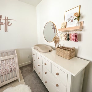 May include: White dresser with nine drawers and wooden knobs. A white changing pad sits on top of the dresser. A wicker basket and a round mirror are also on the dresser. A wooden shelf with a gold metal rod hangs on the wall above the dresser. The shelf holds a framed print with floral artwork and a vase with dried flowers.
