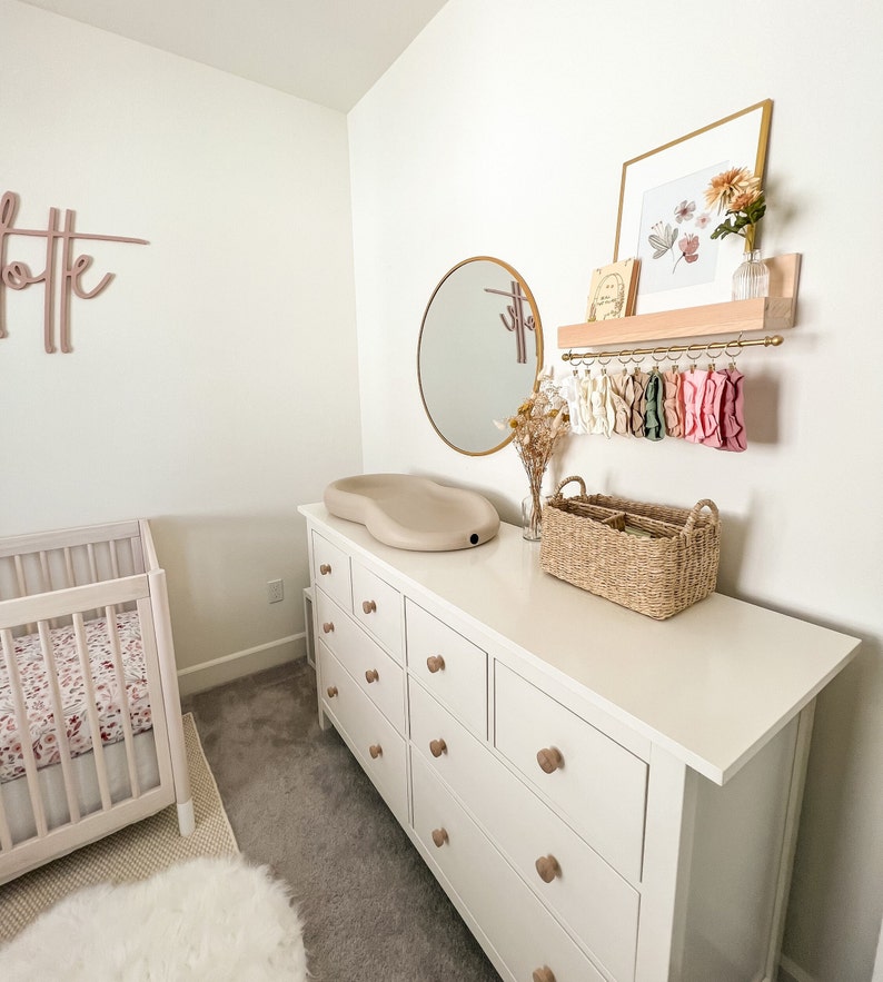 May include: White dresser with nine drawers and wooden knobs. A white changing pad sits on top of the dresser. A wicker basket and a round mirror are also on the dresser. A wooden shelf with a gold metal rod hangs on the wall above the dresser. The shelf holds a framed print with floral artwork and a vase with dried flowers.