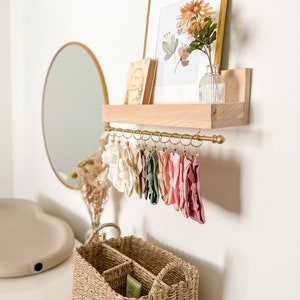 May include: A wooden shelf with a gold metal rod and hooks, holding a collection of colorful headbands. A framed print with a floral design and a small book are on the shelf. A wicker basket with dividers sits on a white surface below the shelf.