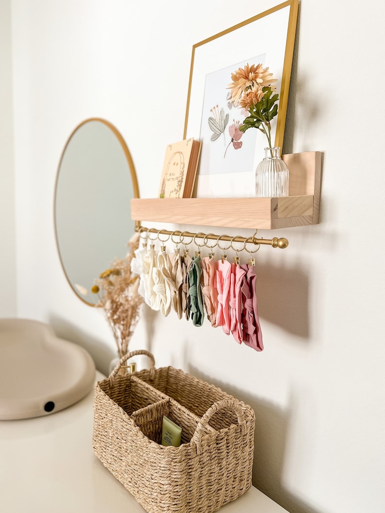 May include: A wooden shelf with a gold metal rod and hooks, holding a collection of colorful headbands. A framed print with a floral design and a small book are on the shelf. A wicker basket with dividers sits on a white surface below the shelf.