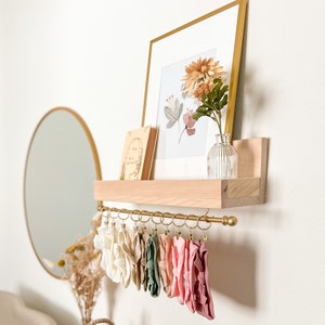 May include: A wooden shelf with a gold metal rod for hanging items. The shelf is decorated with a framed print of flowers and a book. There are several colorful fabric headbands hanging from the rod.