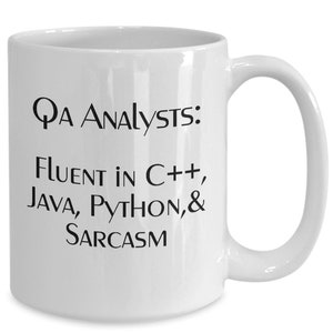 Könnte beinhalten: Weiße Keramik-Kaffeetasse mit schwarzem Text, der "QA Analysts: Fluent in C++, JAVA, Python, & SARCASM" lautet.