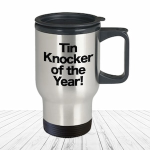 May include: A stainless steel travel mug with a black lid and handle. The mug has the text "Tin Knocker of the Year!" printed on it.