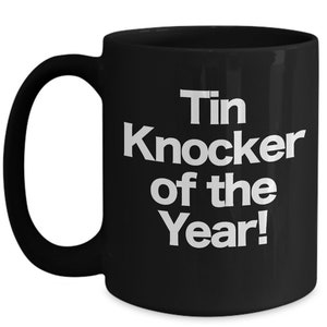 May include: Black coffee mug with white text that reads "Tin Knocker of the Year!"