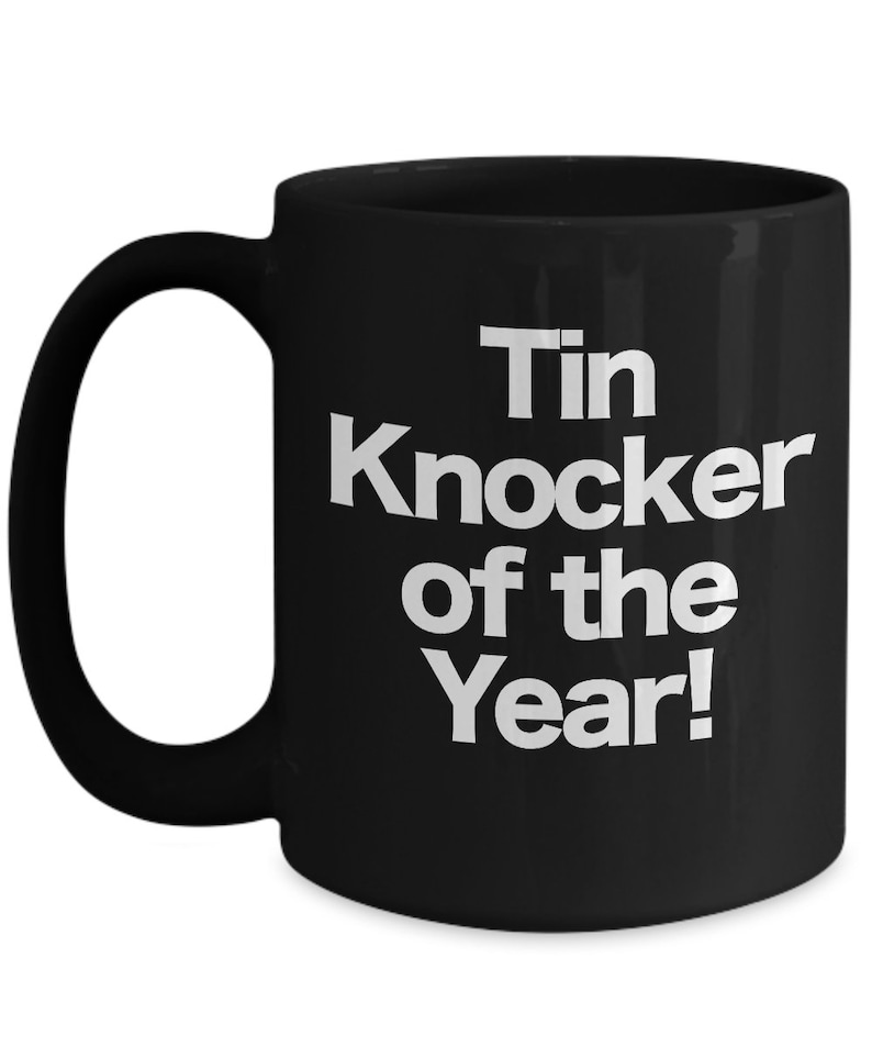 May include: Black coffee mug with white text that reads "Tin Knocker of the Year!"