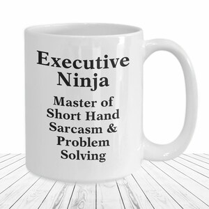 May include: White ceramic coffee mug with black text that reads "Executive Ninja Master of Short Hand Sarcasm & Problem Solving".