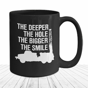 Könnte beinhalten: Schwarze Kaffeetasse mit weißem Text, der "The deeper the hole the bigger the smile" lautet. Die Tasse zeigt außerdem eine weiße Silhouette eines Bohrgerüsts und eines Lastwagens.